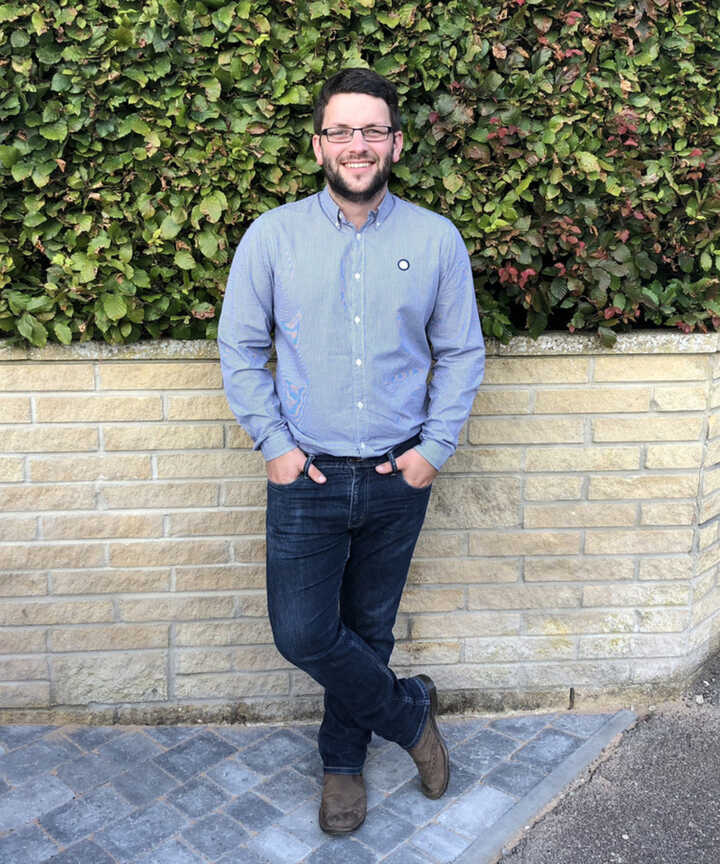 Jim Larcombe stands in a grey shirt and jeans by a stone wall and greenery.